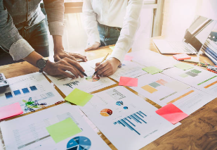 Close-up of a diverse group of professional colleagues collaborating at an office table covered in colorful charts, flowcharts, and project diagrams, working together on project planning.